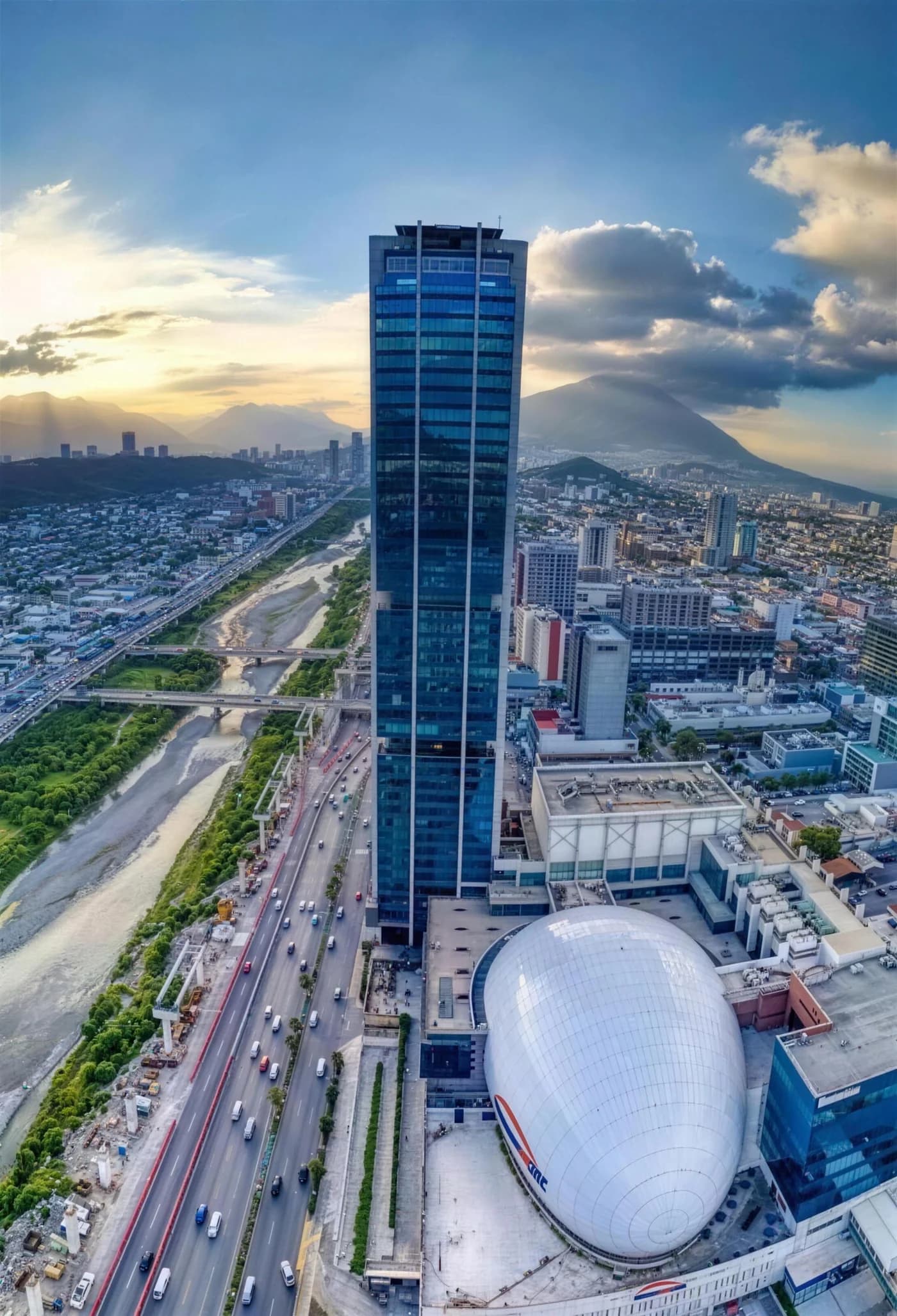 Vista aérea de Pabellón M al atardecer: la torre, el auditorio de cubierta ovalada y la ciudad de Monterrey con el Cerro de la Silla al fondo.
